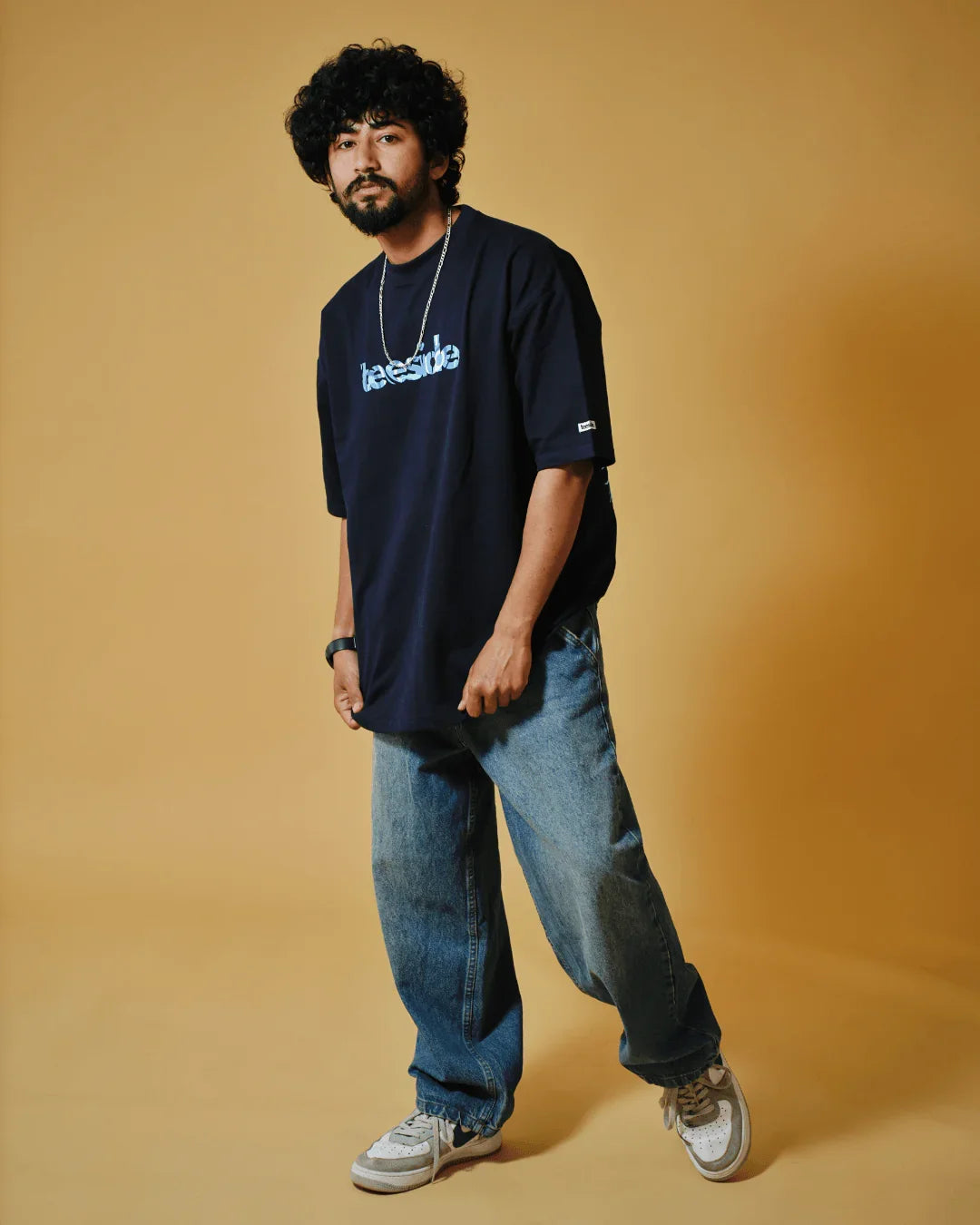Man wearing navy teeside t-shirt, baggy jeans, and sneakers on beige background
