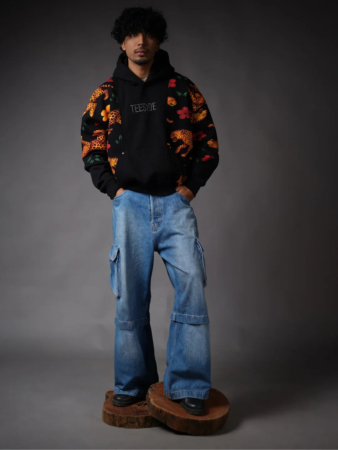 Man wearing TEESIDE black hoodie with leopard and floral print sleeves, wide-leg denim cargo jeans.