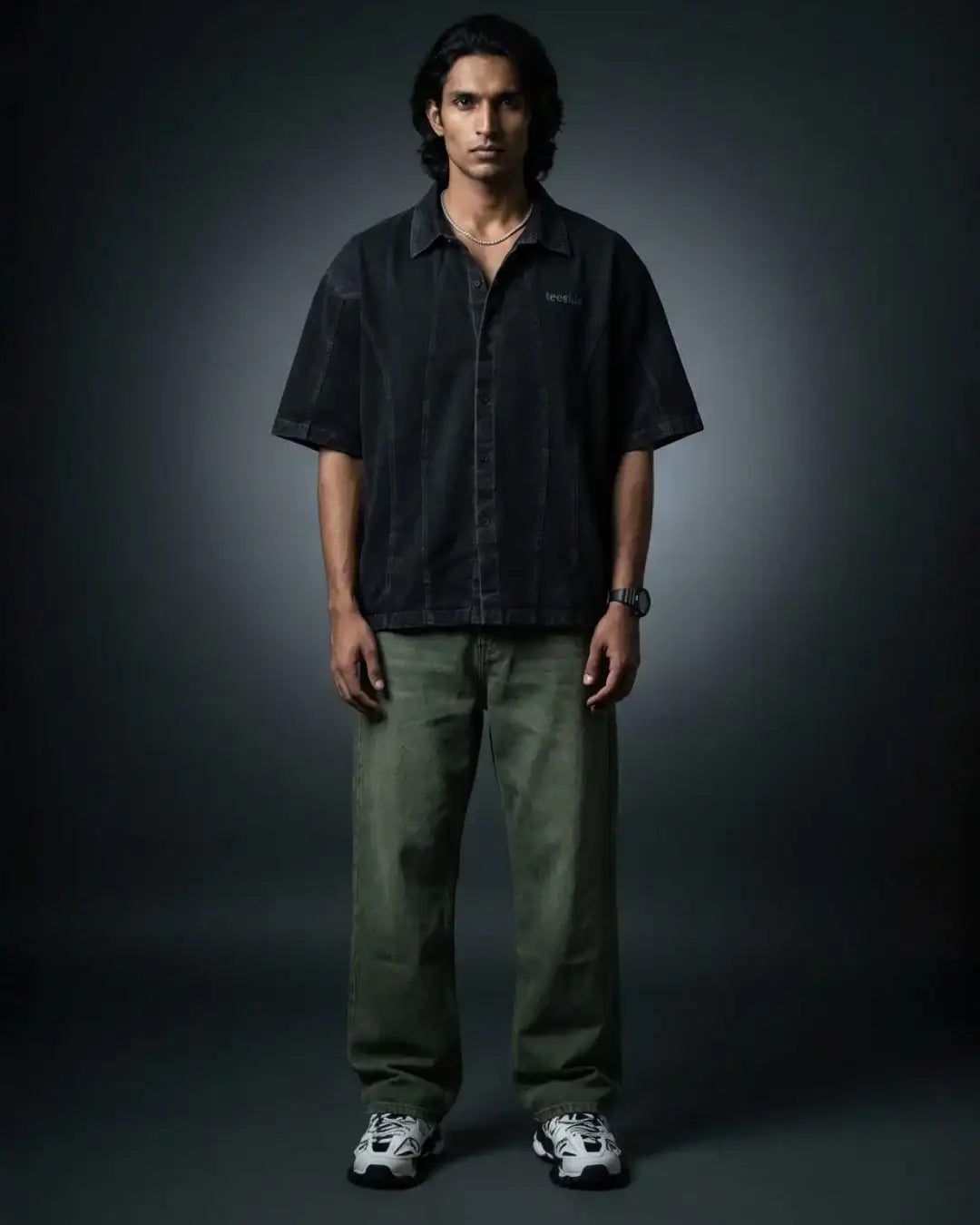 Man wearing a black teeside button-up shirt, green jeans, and sneakers in studio