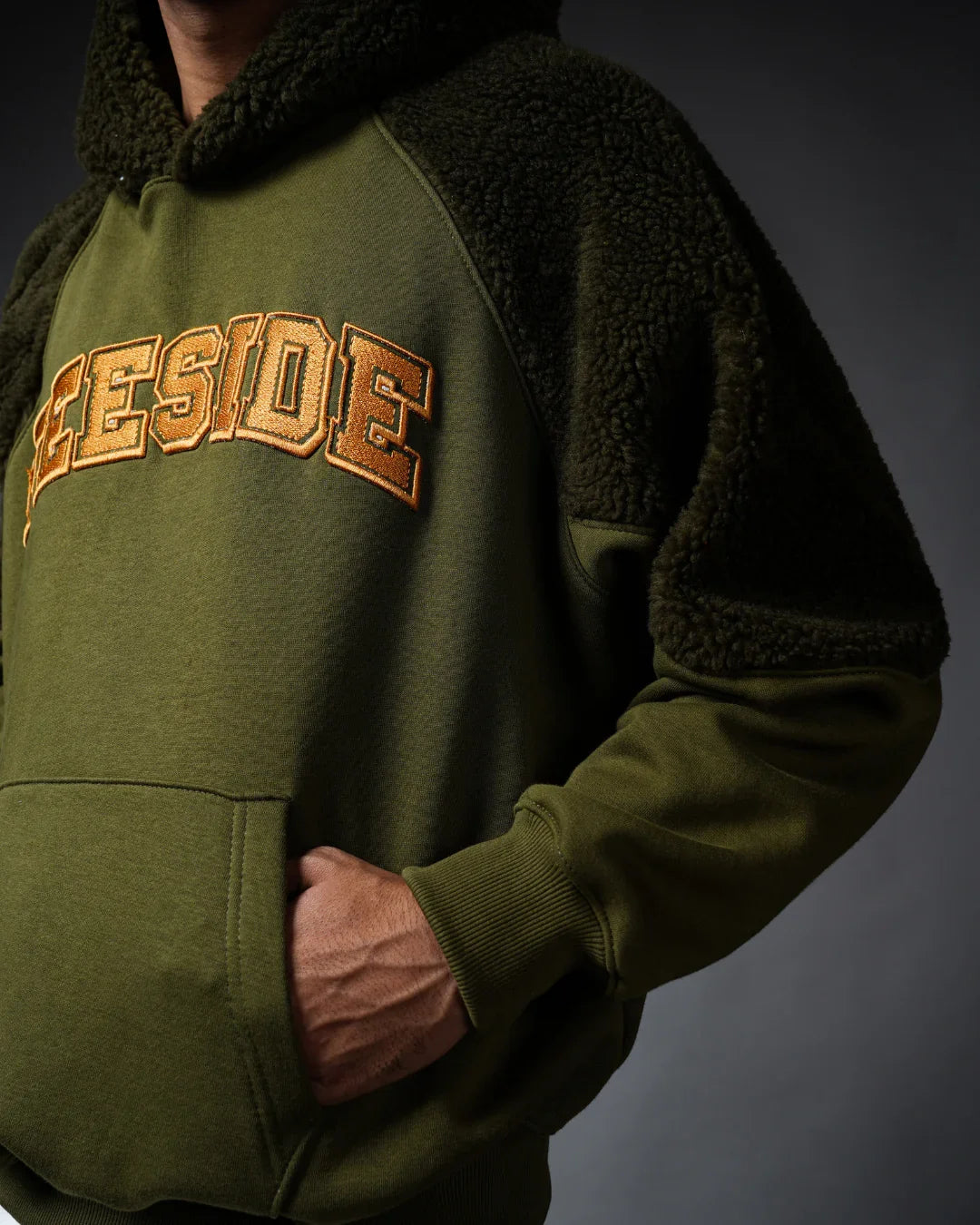 Man wearing an olive green hooded sweatshirt with TEESIDE text and plush textured sleeves