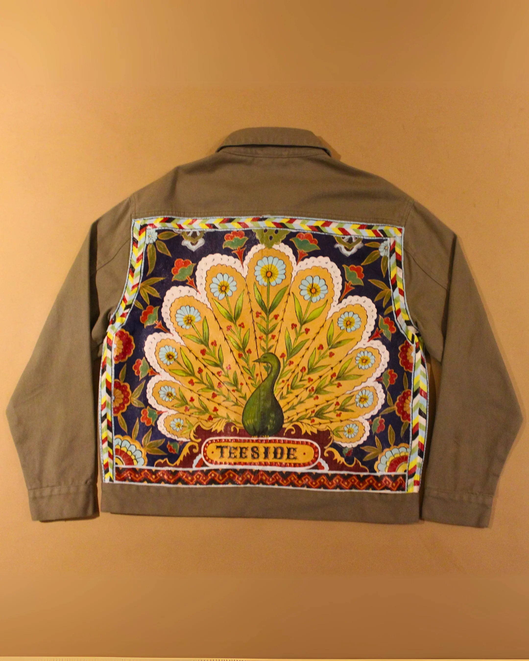 Back view of a khaki jacket with a vibrant peacock and floral design, teeside text on panel