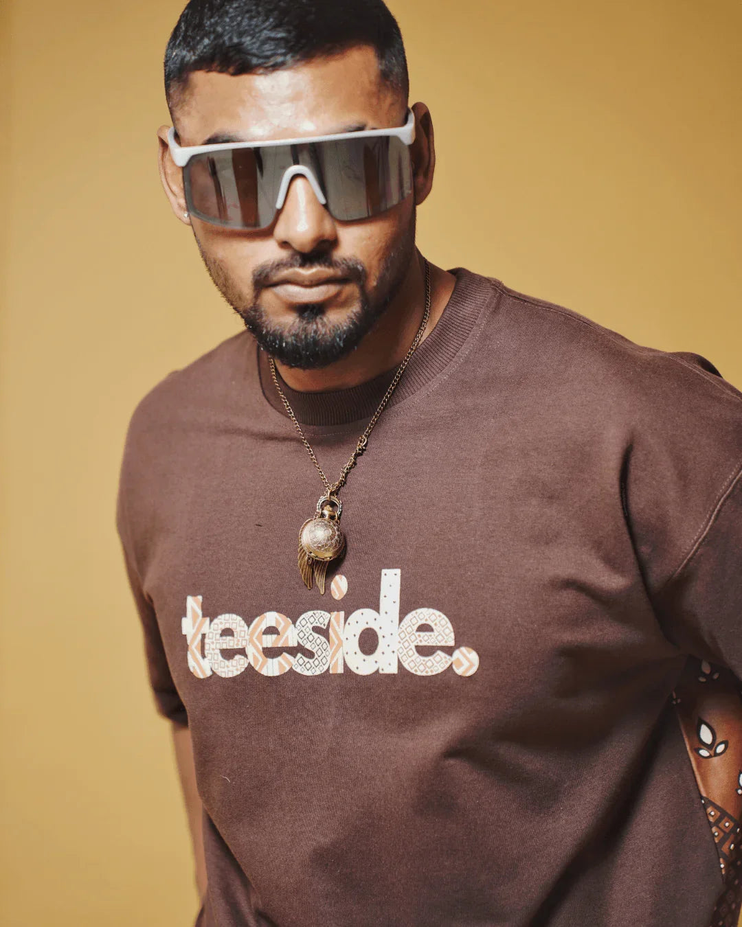 Man wearing brown teeside graphic t-shirt and large sunglasses, posing against yellow background.