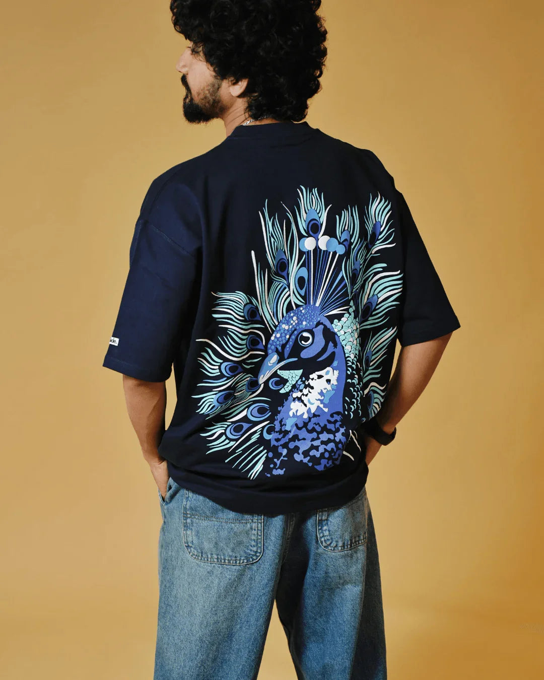 Man wearing dark tee with vibrant peacock graphic by teeside, casual jeans, yellow background
