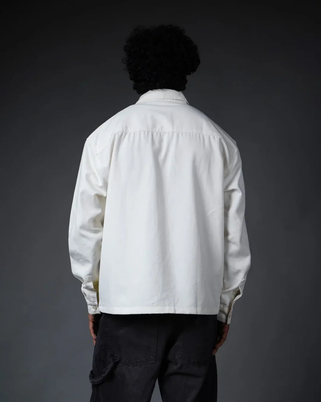 Back view of person wearing a white teeside shirt and black pants against a dark background.